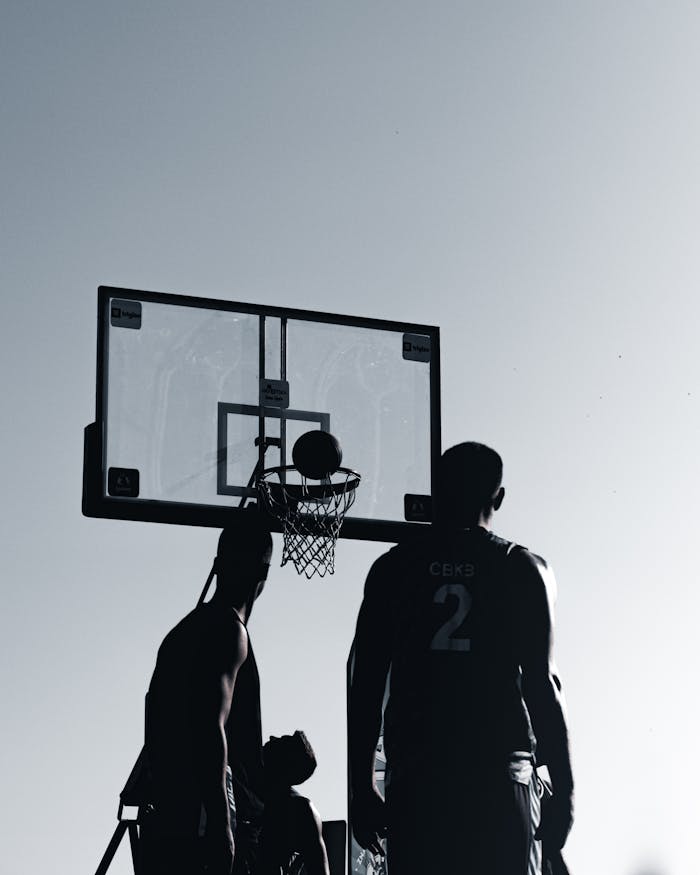 Silhouettes of male basketball players practicing on an outdoor court during daylight.