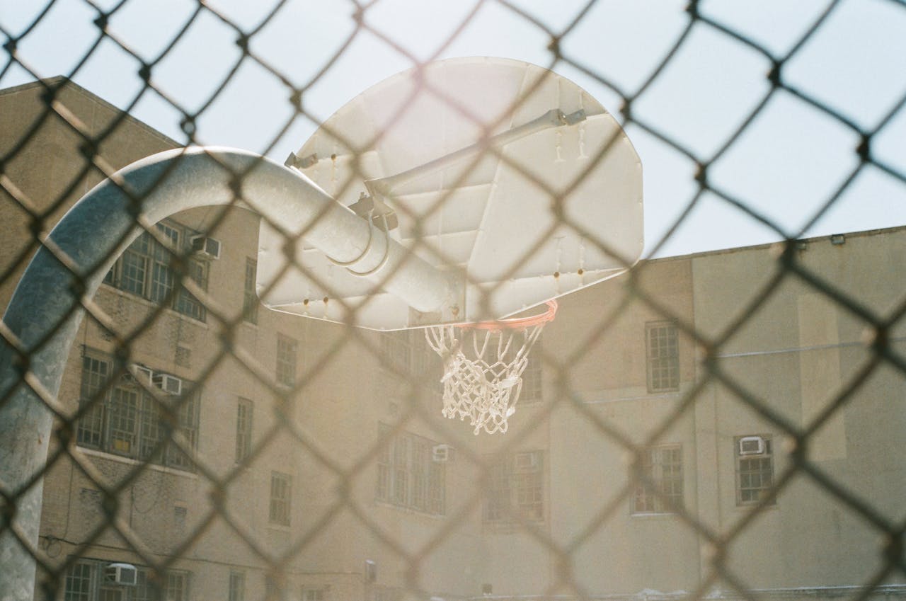 Vintage basketball hoop in urban playground, seen through a chain-link fence on a sunny day.